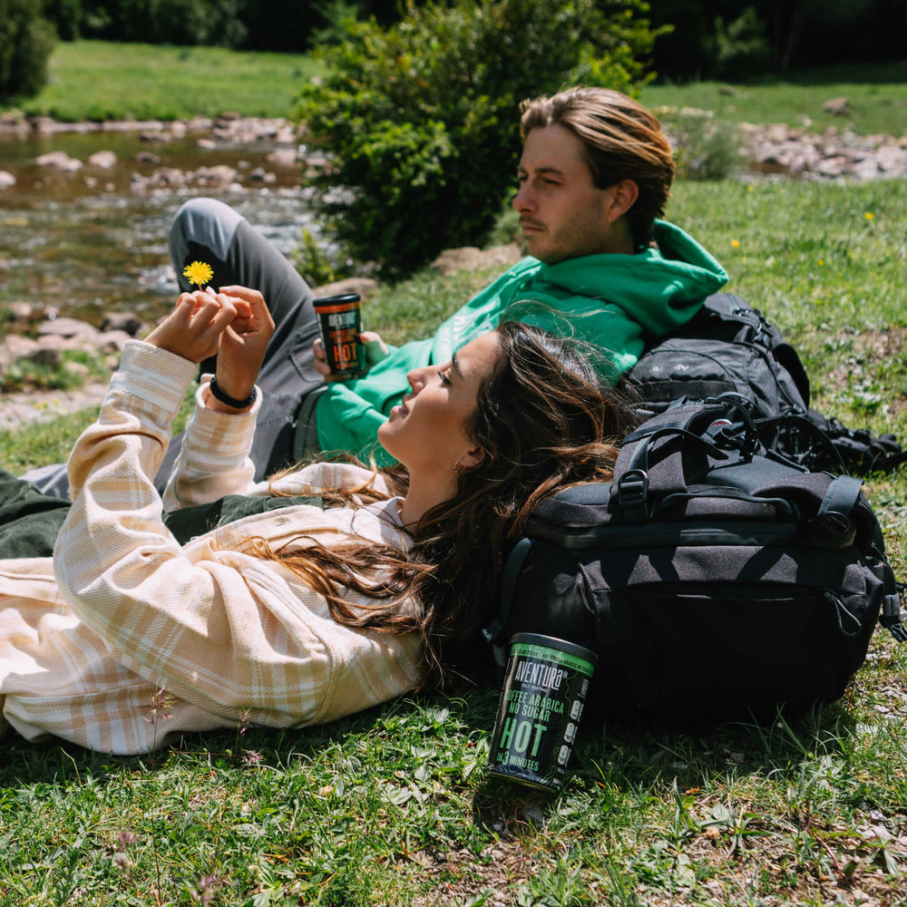 
                  
                    Aventura Self Heating Coffee Arabica No Sugar Hot in 3 Minutes and Aventura Self Heating Cappuccino Caramel next to 2 people on an adventure in nature next to a lake holding a dandelion and resting on hiking backpacks
                  
                